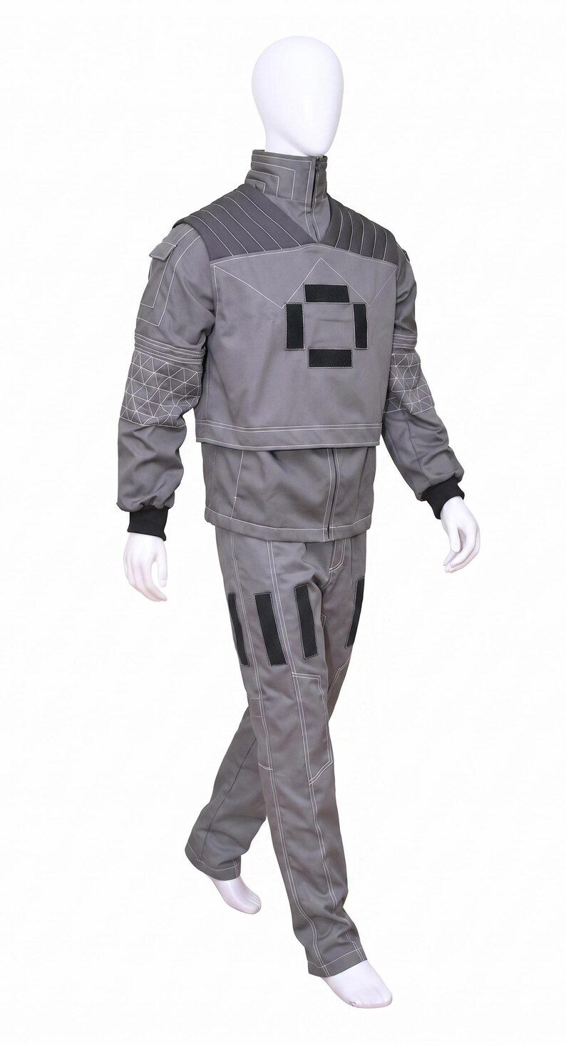 May include: A grey two-piece outfit consisting of a long-sleeved jacket and matching trousers. The jacket features a high collar, black rectangular accents on the chest, and quilted shoulder panels. The trousers have black rectangular patches.