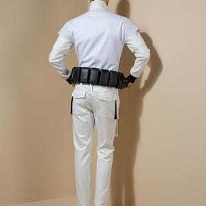 Pu&ograve; includere: Un costume bianco con collo alto, maniche lunghe e cintura nera con tasche. I pantaloni sono bianchi con dettagli neri. La testa e i piedi del manichino sono dorati.
