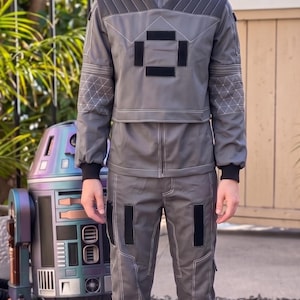 May include: A grey two-piece outfit consisting of a long-sleeved jacket with a high collar, quilted shoulder panels, and geometric stitching on the sleeves, paired with matching trousers featuring black rectangular accents and white contrast stitching.