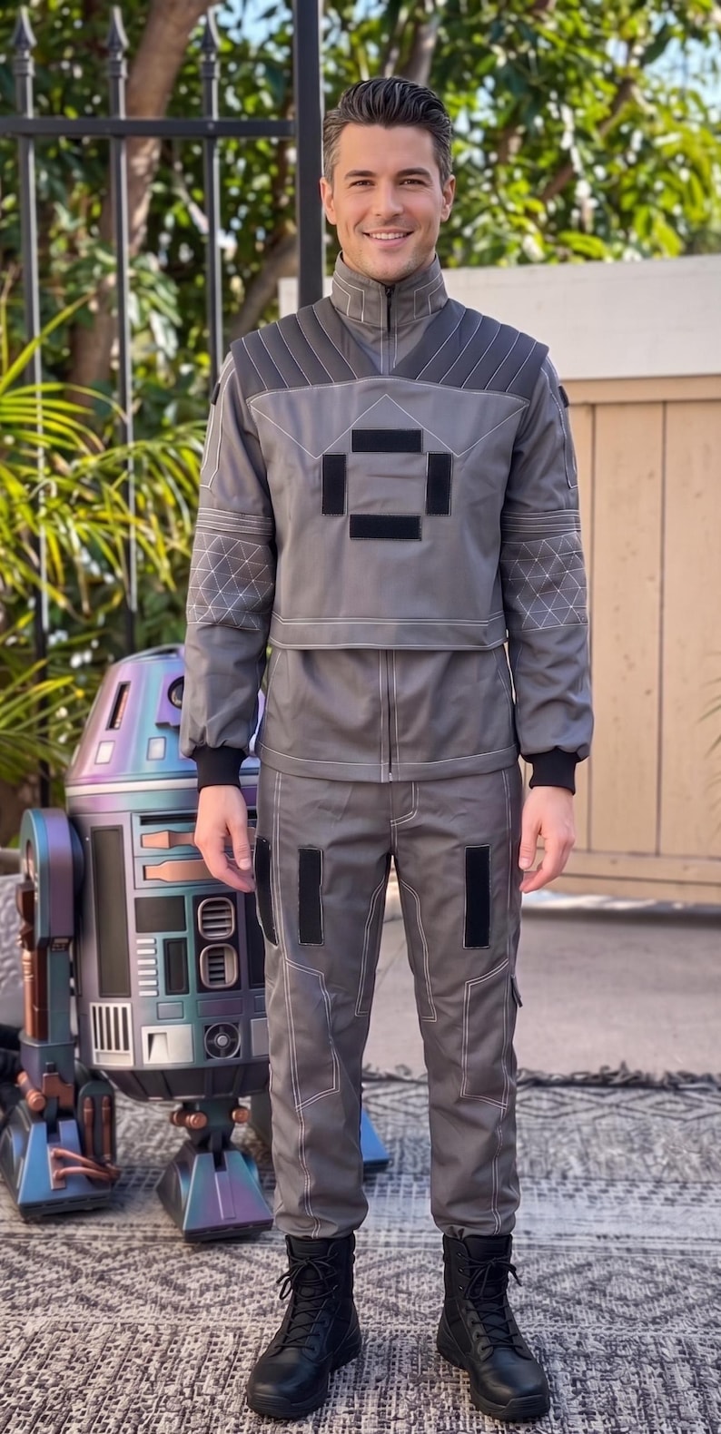 May include: A grey two-piece outfit consisting of a long-sleeved jacket with a high collar, quilted shoulder panels, and geometric stitching on the sleeves, paired with matching trousers featuring black rectangular accents and white contrast stitching.
