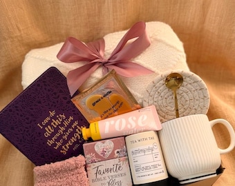 Puede incluir: Una cesta de regalo con una manta blanca atada con una cinta rosa. Contiene un libro morado con letras doradas, un calcetín rosa difuso, una taza blanca y otros artículos de cuidado personal. La cesta está en una bandeja marrón.