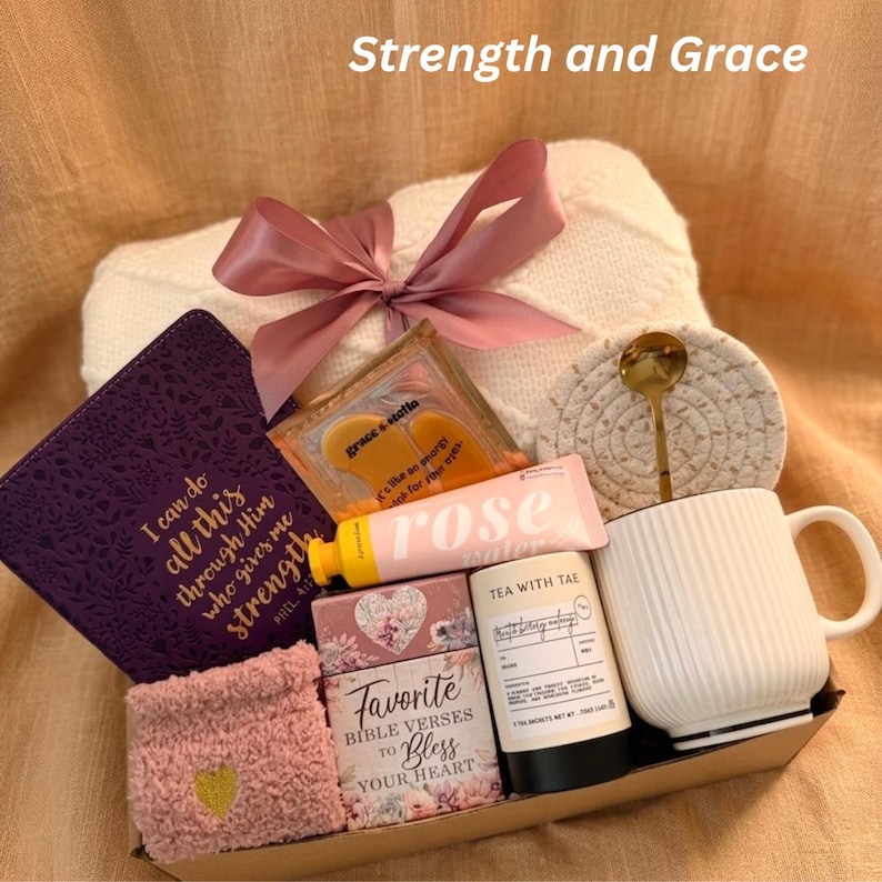 Puede incluir: Una cesta de regalo con varios art&iacute;culos, incluyendo un diario morado con texto dorado, una taza blanca, un tubo rosa de loci&oacute;n "rose" y una peque&ntilde;a caja de "Favorite Bible Verses". El texto "Strength and Grace" es visible.