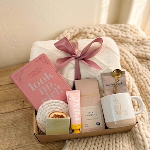 Op de afbeelding: Een cadeaupakket met een roze dagboek met de tekst "look up girl!", een witte deken met een roze lint, een mok, chocolade, handcrème en zeep. De items zijn gerangschikt in een bruine tray.