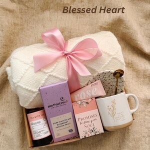 Puede incluir: Una caja de regalo con una manta color crema atada con una cinta rosa. La caja contiene una taza, t&eacute;, chocolate y un plato peque&ntilde;o. La taza tiene un dise&ntilde;o floral y las palabras "Always and Never". El texto "Blessed Heart" est&aacute; en la parte superior.