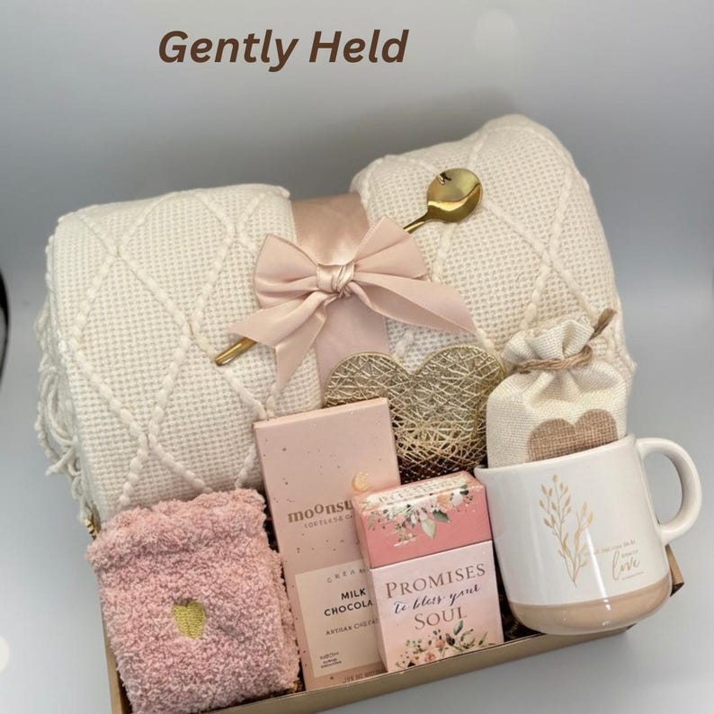 Puede incluir: Una cesta de regalo con una manta color crema, lazo rosa, cuchara dorada, decoraci&oacute;n en forma de coraz&oacute;n, calcetines rosas, barras de chocolate, una taza con la palabra "love" y una bolsita. El texto "Gently Held" est&aacute; en la parte superior.