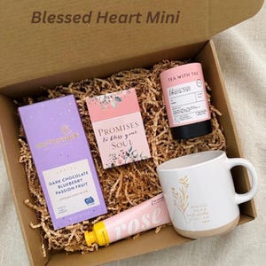Puede incluir: Una caja de regalo "Blessed Heart Mini" llena de una taza blanca con base beige y dise&ntilde;o floral dorado, t&eacute;, chocolate, una tarjeta y loci&oacute;n de rosas. El chocolate es morado y la tarjeta dice "Promises to bless your soul".