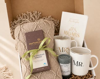 Puede incluir: Una caja de regalo que contiene una manta de punto beige con flecos, una barra de chocolate negro espresso Moonstruck con un lazo verde, una lata pequeña gris de té Tea With Tae, dos tazas blancas con MR. y MRS. y un libro titulado For Couples.
