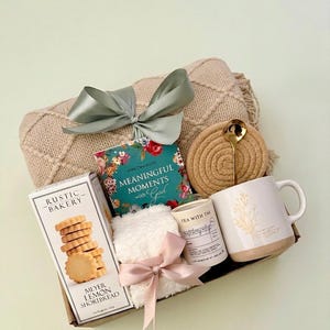 Puede incluir: Una cesta de regalo llena de una manta beige, un libro titulado "Meaningful Moments with God", una taza con un dise&ntilde;o floral, una caja de galletas Meyer Lemon Shortbread, una vela peque&ntilde;a, un posavasos, una cuchara y una peque&ntilde;a toalla blanca con un lazo rosa.