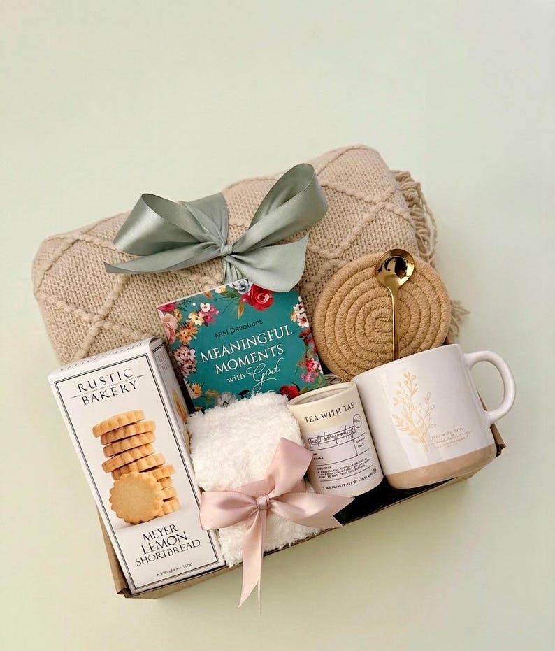Puede incluir: Una cesta de regalo llena de una manta beige, un libro titulado "Meaningful Moments with God", una taza con un dise&ntilde;o floral, una caja de galletas Meyer Lemon Shortbread, una vela peque&ntilde;a, un posavasos, una cuchara y una peque&ntilde;a toalla blanca con un lazo rosa.