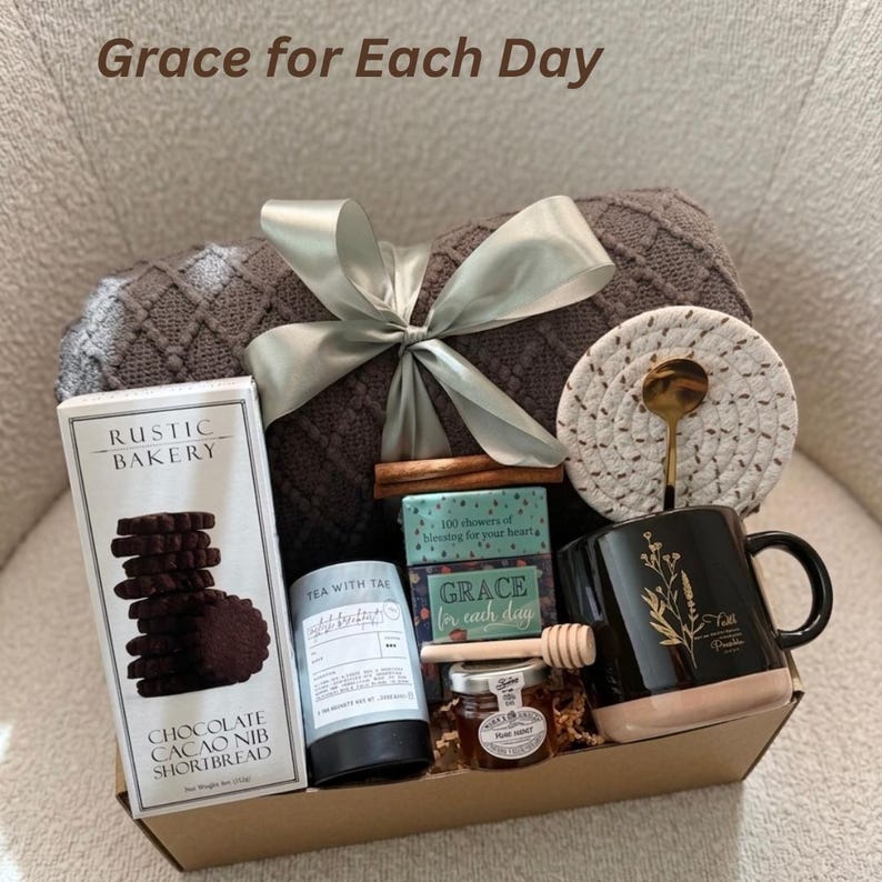 Puede incluir: Una cesta de regalo con una manta gris atada con una cinta plateada, una taza negra con dise&ntilde;o floral dorado, galletas, t&eacute;, miel y un posavasos. El texto "Grace for Each Day" es visible.