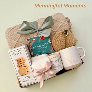 Puede incluir: Una cesta de regalo con una manta beige, un libro titulado "Meaningful Moments with God", una taza, una vela, una caja de galletas de mantequilla de lim&oacute;n y una peque&ntilde;a toalla blanca con un lazo rosa. El texto "Meaningful Moments" est&aacute; en la parte superior.