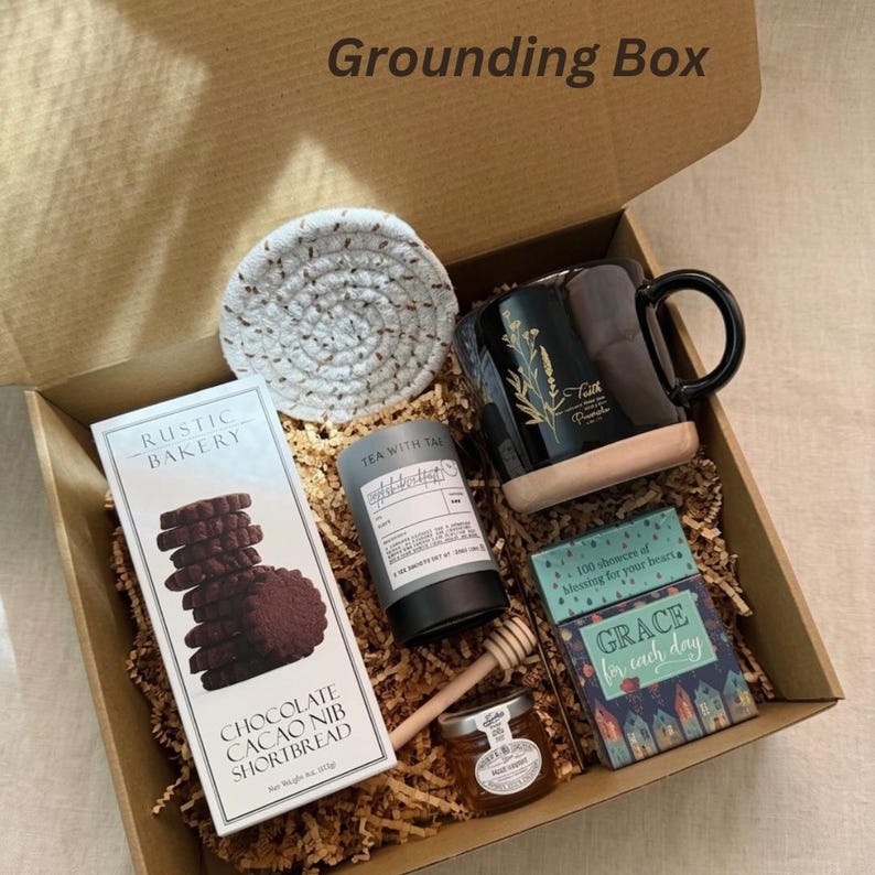 Puede incluir: Una caja de regalo "Grounding Box" llena de una variedad de art&iacute;culos. Incluye una taza negra con dise&ntilde;o floral dorado, galletas de chocolate, t&eacute;, miel, un posavasos y un libro. La caja est&aacute; llena de papel marr&oacute;n.