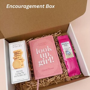 Puede incluir: Una caja de cart&oacute;n abierta, etiquetada como "Encouragement Box", llena de art&iacute;culos. En su interior hay un diario rosa con la frase "look up, girl!", una caja de galletas Meyer Lemon Shortbread y una bolsa de caf&eacute; Strawberry Fields.
