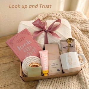Puede incluir: Una caja de regalo con un diario rosa, una taza blanca con un dise&ntilde;o floral, una pastilla de jab&oacute;n, loci&oacute;n con aroma a rosa, una tableta de chocolate, una cuchara dorada y un posavasos. La caja est&aacute; atada con una cinta rosa y contiene el texto "Look up and Trust."