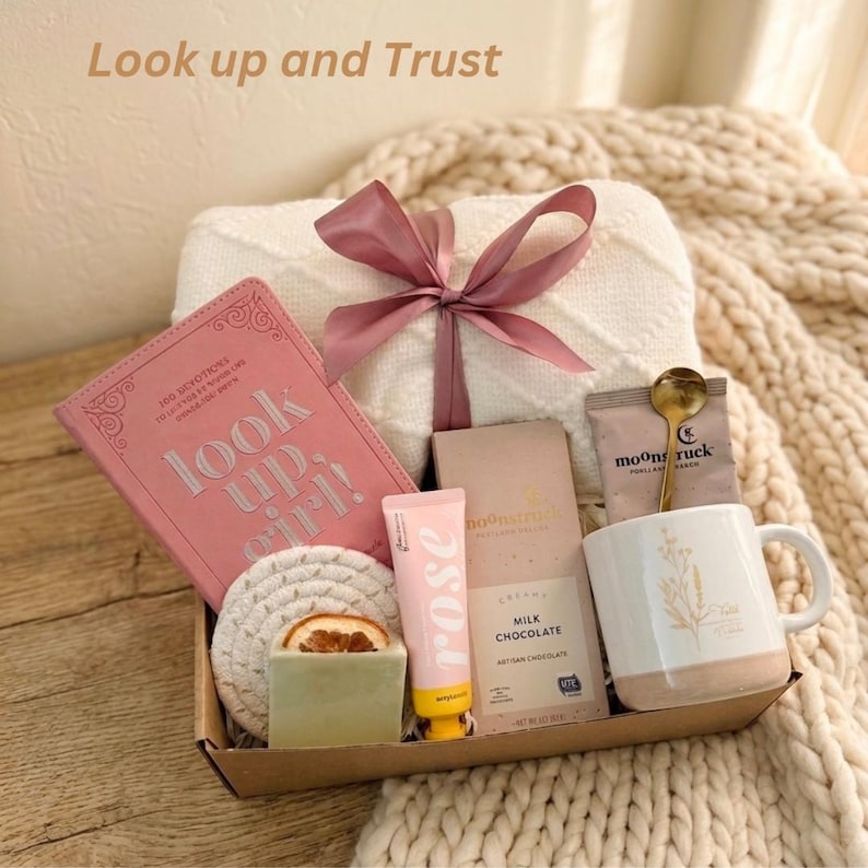 Puede incluir: Una caja de regalo con un diario rosa, una taza blanca con un dise&ntilde;o floral, una pastilla de jab&oacute;n, loci&oacute;n con aroma a rosa, una tableta de chocolate, una cuchara dorada y un posavasos. La caja est&aacute; atada con una cinta rosa y contiene el texto "Look up and Trust."