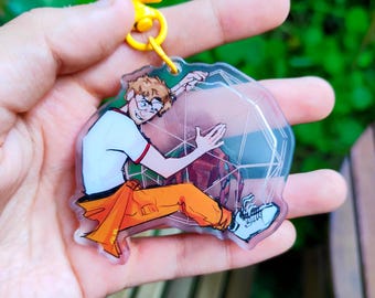 May include: A clear acrylic keychain featuring a cartoon character with brown hair and glasses, wearing a white t-shirt and orange pants, interacting with a white geometric wireframe sphere. The keychain has a yellow star-shaped plastic clasp.