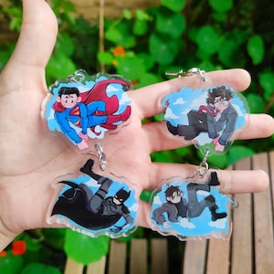May include: Four acrylic keychains featuring comic book characters Superman, Batman, and two other figures. Each keychain has a cartoon illustration against a blue sky background. The keychains are held in a hand against a backdrop of green foliage.