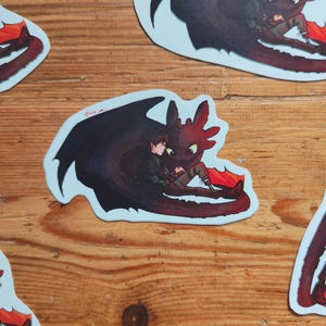 May include: Multiple stickers depicting a cartoon dragon and a person. The dragon is black and red, and the person is dressed in green and brown. The stickers are set against a light blue backdrop.