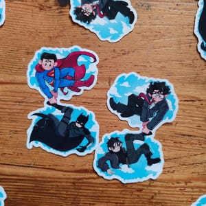 May include: A collection of stickers featuring comic book characters Superman, Batman, and others. Each sticker depicts a character in a dynamic pose against a blue sky and cloud background. The stickers are on a wooden surface.