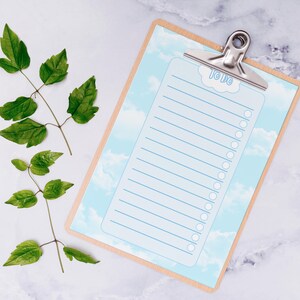 Puede incluir: Un portapapeles de madera con una lista de tareas pendientes azul claro, con un diseño de nubes. El portapapeles tiene un clip plateado en la parte superior y está acompañado de hojas verdes. El texto "TO DO" es visible en la parte superior de la lista.