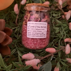 May include: A glass jar filled with pink bath salts and dried rose petals, topped with a cork lid. The label reads "Garden of Venus Bath Salts". The jar is surrounded by greenery and decorative elements, creating a spa-like aesthetic.