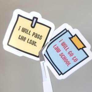 May include: Two stickers with white borders and the text "i will pass the lsat" and "i will go to law school". The first sticker is yellow with a black border and a silver clip. The second sticker is light blue with a black border and a piece of yellow tape.