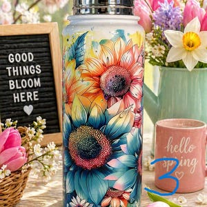 K&ouml;nnte beinhalten: Eine Edelstahl-Wasserflasche mit einem lebendigen Blumendesign mit Sonnenblumen in Orange-, Blau- und Gelbt&ouml;nen. Die Flasche hat einen silbernen Deckel und ist mit einem farbenfrohen, k&uuml;nstlerischen Muster verziert. Der Text "Good Things Bloom Here" ist auf einem kleinen Schild zu sehen.