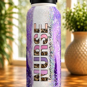 May include: A stainless steel water bottle with a black handle and lid. The bottle features a design with the word "NURSE" in pink and leopard print, with the words "love inspire heal" in cursive. The background has a purple and white glitter design.