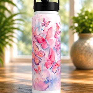 May include: A white water bottle with a watercolour design of pink and purple butterflies. The bottle has a black handle and lid. The background has a watercolour effect with pink, purple, and blue hues. The bottle is on a wooden surface.