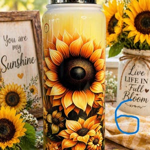 May include: A stainless steel water bottle with a vibrant sunflower design. The bottle features a large, detailed sunflower in shades of yellow, orange, and brown. The background is a gradient of yellow. The bottle has a silver lid and is surrounded by sunflowers.