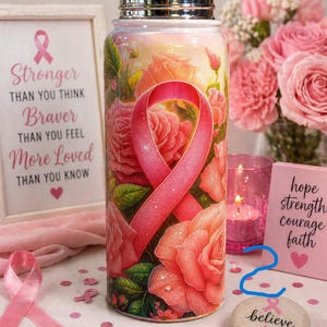 May include: A stainless steel water bottle with a pink ribbon and rose design. The bottle has a silver lid and is surrounded by pink roses, a framed print with text, and a stone that says "believe".