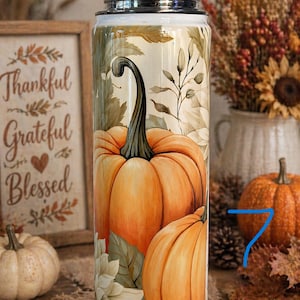 May include: A stainless steel tumbler featuring a vibrant orange pumpkin design with green leaves and a black stem. The tumbler has a silver lid and is decorated with fall foliage. The words "Thankful, Grateful, Blessed" are visible in the background.