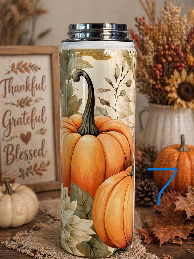 May include: A stainless steel tumbler featuring a vibrant orange pumpkin design with green leaves and a black stem. The tumbler has a silver lid and is decorated with fall foliage. The words "Thankful, Grateful, Blessed" are visible in the background.