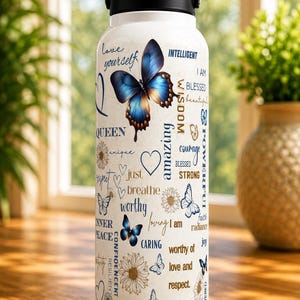 Puede incluir: Botella de agua blanca con asa y tapa negras, decorada con mariposas azules y palabras inspiradoras como "&aacute;mate" e "imparable". La botella tiene un asa y una tapa negras.