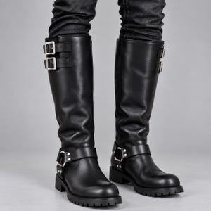 May include: A pair of black knee-high boots featuring two silver-toned buckles on the upper shaft and a harness strap with a silver-toned metal ring at the ankle. The boots have a rounded toe, a low block heel, and a thick, lugged rubber outsole.