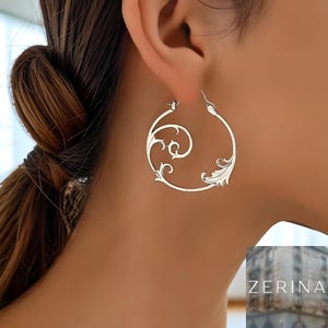 May include: Silver hoop earrings with an openwork floral design. The earrings have a swirling pattern with leaf-like details. The word "ZERINA" is visible in the bottom right corner.