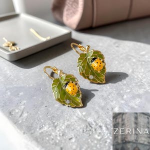 Könnte beinhalten: Goldfarbene Blätterohrringe mit Marienkäfer-Design. Die Ohrringe zeigen ein grünes Blatt mit einem gelb-schwarzen Marienkäfer und einem kleinen, klaren Kristall. Auf einer grauen Oberfläche.