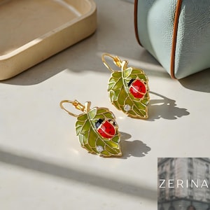 May include: Gold-coloured leaf-shaped earrings with a ladybird design. The earrings feature a green enamel leaf with a red and black ladybird and a small crystal accent. The earrings are on a white surface.