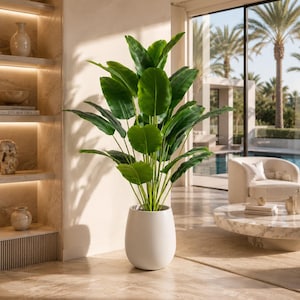 Puede incluir: Una planta de plátano artificial con hojas verdes grandes, en una maceta blanca en forma de huevo. La planta está en el interior, cerca de una ventana con vistas a palmeras y una piscina. La habitación tiene un diseño moderno y minimalista. La maceta mide unos 60 cm de alto.