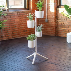 May include: A white vertical plant stand with a tripod base featuring four cylindrical white pots arranged at varying heights. Each pot contains a green houseplant, creating a tiered display structure for indoor greenery and botanical arrangements.