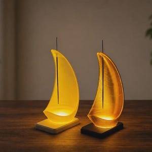 May include: Two sailboat-shaped incense holders with integrated LED lights. One holder is translucent yellow on a white base, and the other is a metallic gold on a black base. Each features a thin, dark incense stick standing upright in the centre.