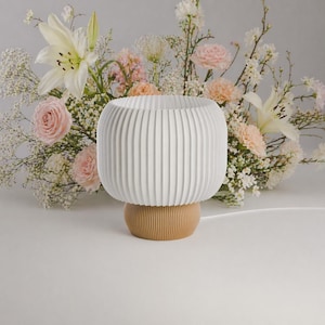 Op de afbeelding: Een witte, geribbelde tafellamp met een ronde kap en een lichtbruine voet. De lamp staat tegen een achtergrond van verse bloemen, waaronder witte lelies en roze rozen, wat een zachte, uitnodigende esthetiek creëert.