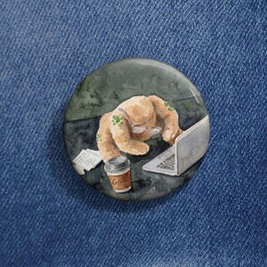 May include: A round button with a watercolor illustration of a furry creature wearing glasses and working on a laptop. A coffee cup and papers are on the desk. The button is set against a blue denim background.