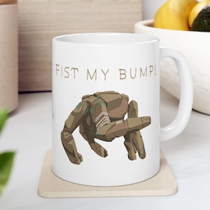 May include: White ceramic mug with the text "FIST MY BUMP!" above a brown, robotic crab illustration. The crab has green and white accents. The mug has a curved handle and is sitting on a beige coaster.