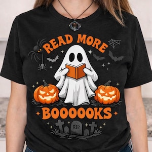 Op de afbeelding: Een zwart t-shirt met korte mouwen en een afbeelding van een witte geest die een oranje boek leest. De tekst luidt READ MORE BOOOOOKS. Het ontwerp bevat twee oranje pompoenen, een spin, vleermuizen, een spinnenweb en een grafsteen met RIP.