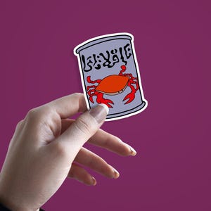 May include: A sticker featuring a cartoon crab on a can. The can is gray with white trim and has text in a foreign language. The crab is orange with red claws. The sticker is held against a purple background.