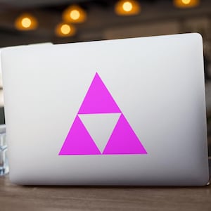 May include: A silver laptop with a bright pink and white triangle design on the lid. The design is a stylized geometric shape. The laptop is on a wooden table, with a glass of water visible in the foreground.