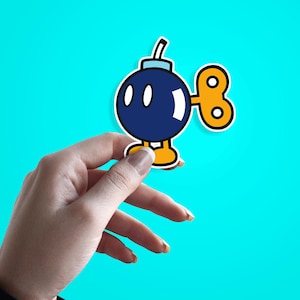 Puede incluir: Una pegatina con un personaje de bomba de dibujos animados con un cuerpo azul oscuro, ojos blancos y una llave de cuerda amarilla. La bomba tiene un contorno blanco y está sobre un fondo turquesa brillante. La pegatina está sujeta por una mano.