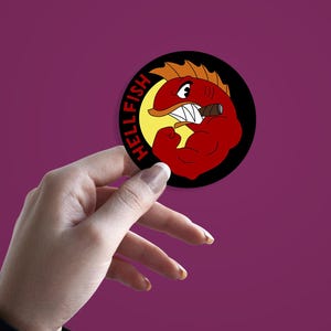 May include: A round sticker with a cartoon illustration of a red fish with orange fins, a cigar, and flexing arm. The sticker has the word "HELLFISH" in red letters on a black background.