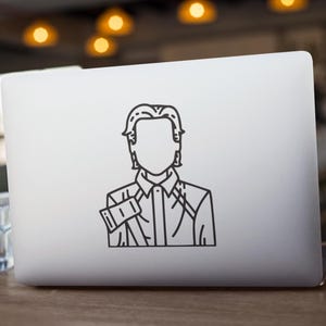 May include: A white laptop displaying a black line art sticker of a person. The sticker shows a person in a collared shirt and jacket. The laptop is on a wooden surface, with a glass of water in the foreground.